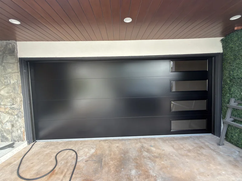 Dark flush-panel garage door with four side windows beneath a wood-toned ceiling.